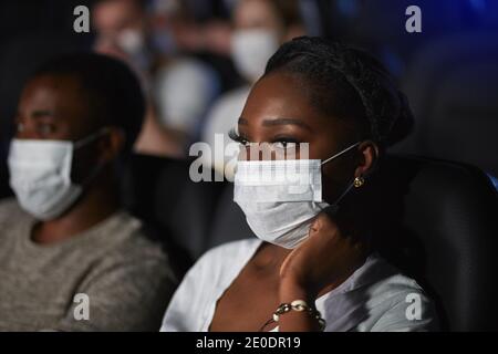 Nahaufnahme einer afrikanischen Frau, die während der Weltpandemie Film genießt. Selektiver Fokus des jungen Paares, das Film im Kino sieht, in bequemen schwarzen Sitzen sitzt, weiße Gesichtsmasken trägt. Stockfoto