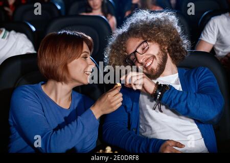 Glückliches kaukasisches Paar, das gemeinsam Komödie im Kino genießt und lächelt. Junge emotionale kaukasische Frau beim Film geben Popcorn Freund mit Afro-Frisur im Kino. Stockfoto