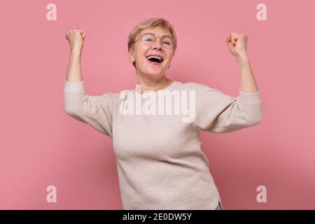 Überglücklich reife alte Frau heben Hände Triumph. Aufgeregt ältere weibliche Blick auf Kamera schreien ja euphorisch fühlen. Studio auf rosa Wand aufgenommen. Stockfoto
