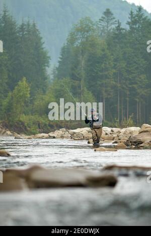 Reifer Mann in Mütze, Brille und spezielle Uniform Angeln mit Spinnen zwischen Bergen. Aktivitäten im Freien in der Nähe von Wasser. Konzept der Fischerei. Stockfoto