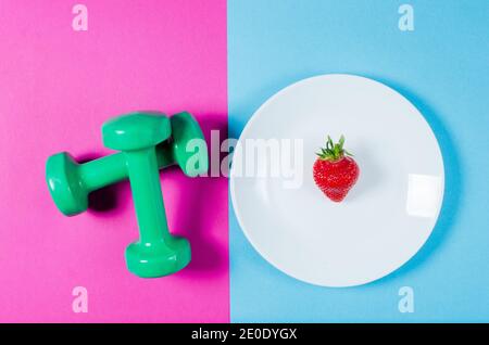 Erdbeere und Sportgewichte liegen auf einem rosa und blauen Hintergrund. Das Konzept der richtigen Ernährung. Gesunder Lebensstil. Ernährung Stockfoto