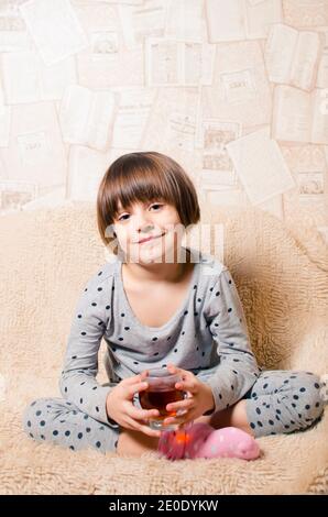 Ein kleines lächelndes Mädchen mit blauen Augen hält ein Glas mit frisch gepresstem Saft in der Hand. Kinderportrait. Feder. Vitamine Stockfoto