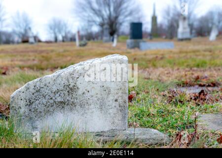 Der Boden eines alten, gebrochenen Grabmarkers. Der Stein ist aus Marmor und gebrochen. Nahaufnahme mit niedrigem Winkel. Andere Gräber im Hintergrund, bedecktem Himmel. Stockfoto