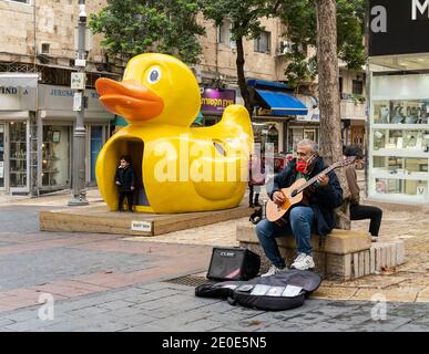 Jerusalem, Israel - 17. Dezember 2020: Ein Mann spielt Gitarre auf einer Jerusalemer Straße, während Kinder um eine Statue einer riesigen Gummiente spielen. Stockfoto