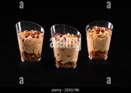 Nahaufnahme von köstlichen süßen Dessert in drei Gläsern in Reihe isoliert auf schwarzem Hintergrund. Leckere frische Parfait mit Müsli, kleine braune Karamellkugeln auf der Oberseite und Schokolade Schlagsahne. Stockfoto