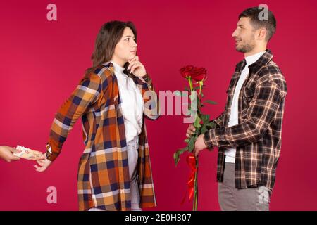 Junger Mann mit roten Rosen, der vor einer Frau steht, die von einem anderen Mann Geld hinter sich nimmt. Roter Hintergrund. Hochwertige Fotos Stockfoto