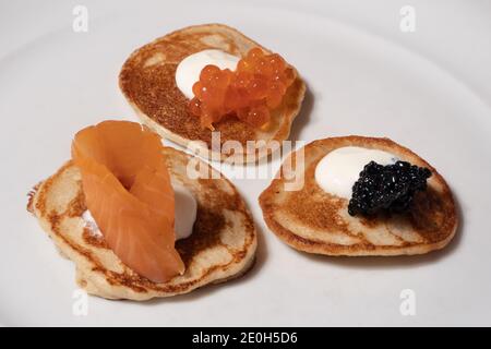 Gemischte Blinis mit geräuchertem Lachs, Rad und weißem Kaviar garniert mit Sour Cream auf einem weißen Teller Stockfoto