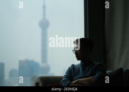 Mitten in der Aufnahme eines jungen asiatischen Mannes, der in Gedanken am Fenster saß, verschwimmte ein modernes Wahrzeichen aus dem Fenster Stockfoto