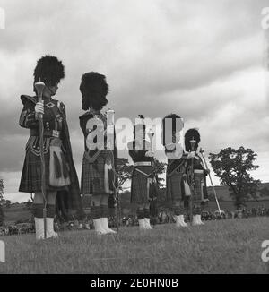 Sechziger Jahre, historisch, draußen bei den Highland Games, Schottland, stehen fünf Drum-Majors in ihren Outfits und mit Maces an, die zur Leitung ihrer schottischen Pfeifenbands verwendet werden, die im 19. Jahrhundert in den schottischen Regimenten der britischen Armee begannen. Historisch gesehen hätte ein Schlagzeugmajor eine leitende Position in der britischen Armee inne. Er war der Leiter des Regiments und wäre ein Anführer der Männer gewesen. Stockfoto