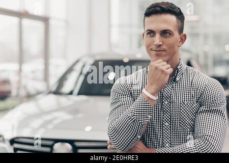 Der junge Mann schaut nachdenklich weg, reibt sich das Kinn und steht vor einem neuen Auto im Autohaus. Schöner Mann, der über den Kauf eines n nachdenkt Stockfoto