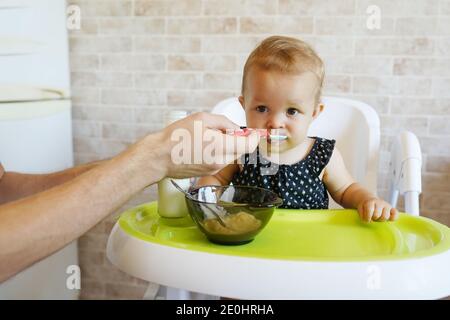 Lustige Kind Mädchen essen aus dem Löffel. Frau Fütterung kleines Baby sitzt in Babystuhl in der Küche. Stockfoto