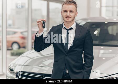 Erfolgreicher Geschäftsmann steht vor einem neuen Auto im Autohaus, hält den Autoschlüssel. Männlicher Kunde posiert in der Nähe seines neuen Autos. Autobesitzer Urlaub Stockfoto