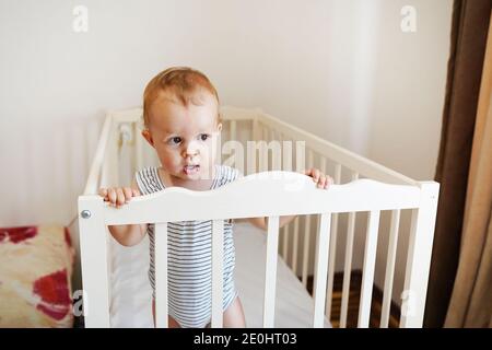 Niedliches Baby steht in einem weißen runden Bett. Weiße Kinderkrippe für Kinder. Kleines Mädchen, das lernt, in seiner Krippe zu stehen. Stockfoto