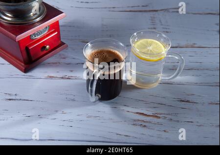 Kaffeemühle mit einer Tasse Kaffee und Wasser mit Zitrone auf einem hellen strukturierten Hintergrund. Stockfoto