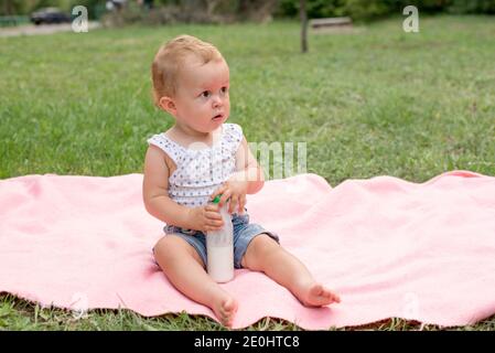 Baby hält Flasche mit Milch oder Kefir, Kind im Park auf grünem Gras. Gesundes Essen für Kinder Stockfoto
