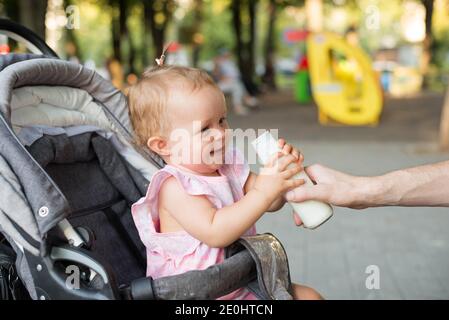 Vater gibt Babymilch oder Kefir, Kind im Kinderwagen im Freien im Park. Ernährung Lebensmittel für Kinder Stockfoto