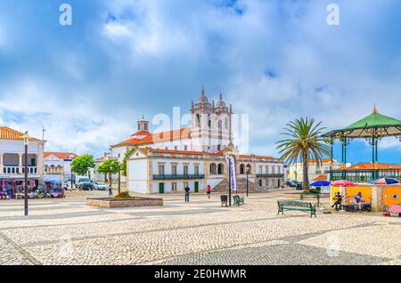 Nazare, Portugal, 22. Juni 2017: Heiligtum unserer Lieben Frau von Nazare katholische Kirche und Straßenmizianer in der Nähe von Pavillon auf gepflasterten Platz mit Palmen auf Sitio Hügel Stockfoto