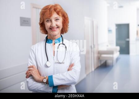 Fröhliche ältere Ärztin lächelt zur Kamera. Freundliche Praktizierende posiert selbstbewusst in ihrer Klinik Stockfoto