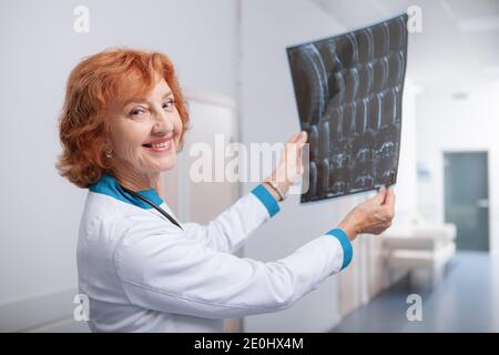 Freundliche Onkologin lächelt zur Kamera und hält die MRT-Untersuchung eines Patienten. Älterer Arzt, der die MRT-Untersuchung eines Patienten untersucht Stockfoto