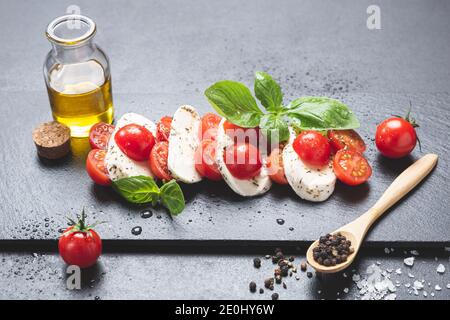 Mozzareela, Tomate und Basilikum, auf Tafel auf schwarzem Hintergrund, mit nativem Olivenöl extra, Salz und Pfeffer. Mediterrane Ernährung und gesunde Ernährung Stockfoto