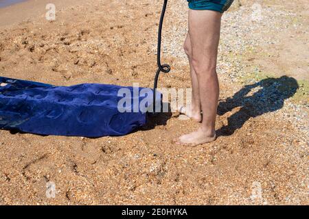 Ein Mann in Badehosen pumpt eine Gummimatratze auf den Sand, an der Küste. Schwimmen auf aufblasbaren Booten. Spaß im Sommer Stockfoto