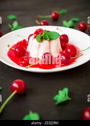 Panna Cotta mit Kokosnussmilch, Kirschsirup und Beeren auf weißem Teller auf dunklem Hintergrund, vegetarisches Dessert Stockfoto