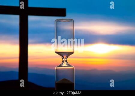 Hourglass hat die Zählung der Zeit auf dem Hintergrund des Sonnenuntergangs und ein Holzkreuz beendet. Ende der Lebensdauer. Der Wert der Zeit im Leben. Stockfoto