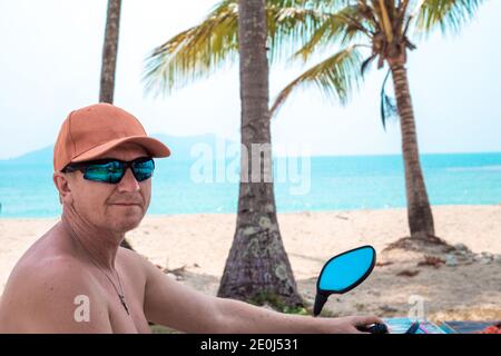 Ein Mann mit Mütze und Brille sitzt auf einem Motorrad, an der Küste mit Palmen. Ruhe auf der tropischen Insel. Fahrradverleih für Reisen Stockfoto