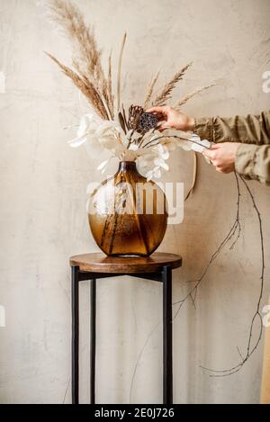 Frau Dekoration Haus mit einer Zusammensetzung von getrockneten Blumen und Kräuter in einer Glasvase auf beigefarbenem Wandhintergrund Stockfoto