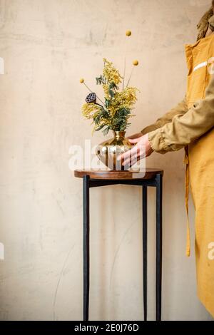 Weibliche Dekokartierung nach Hause mit Komposition aus frischen Blumen und Kräutern in einer goldenen Vase auf einem beigen Wandhintergrund. Bouquet von Acacia, Dealbata, Mimosa, nelumbo und Craspedia Stockfoto