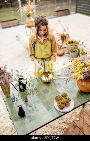 Junge Frau macht Kompositionen aus getrockneten und frischen Blumen und Kräutern in der Werkstatt im Freien. Floristin, Gärtnerin oder Dekorateurin, die Blumendekoration komponiert Stockfoto