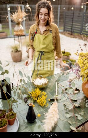 Junge Frau macht Kompositionen aus getrockneten und frischen Blumen und Kräutern in der Werkstatt im Freien. Floristin, Gärtnerin oder Dekorateurin, die Blumendekoration komponiert Stockfoto