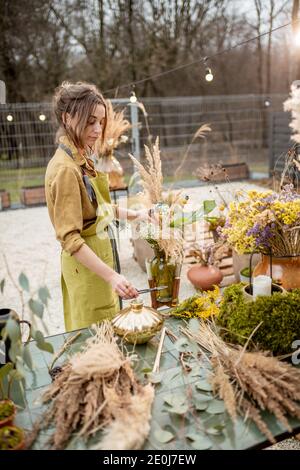 Junge Frau macht Kompositionen aus getrockneten und frischen Blumen und Kräutern in der Werkstatt im Freien. Floristin, Gärtnerin oder Dekorateurin, die Blumendekoration komponiert Stockfoto