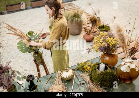 Junge Frau macht Kompositionen aus getrockneten und frischen Blumen und Kräutern in der Werkstatt im Freien. Floristin, Gärtnerin oder Dekorateurin, die Blumendekoration komponiert Stockfoto