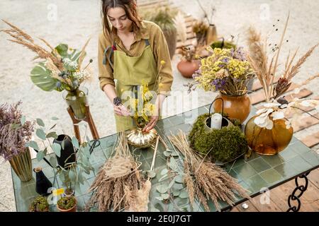 Junge Frau macht Kompositionen aus getrockneten und frischen Blumen und Kräutern in der Werkstatt im Freien. Floristin, Gärtnerin oder Dekorateurin, die Blumendekoration komponiert Stockfoto