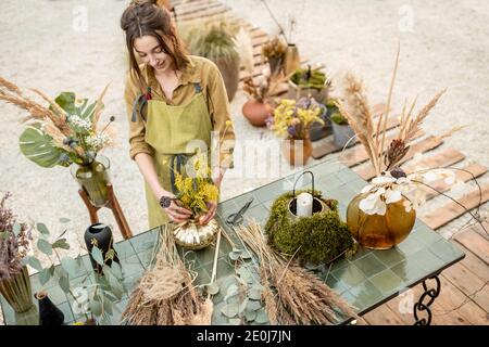 Portrait einer fröhlichen Frau, die in der Werkstatt im Freien Kompositionen aus getrockneten und frischen Blumen und Kräutern macht. Floristin, Gärtner oder Dekorateur Beruf oder Hobby-Konzept Stockfoto