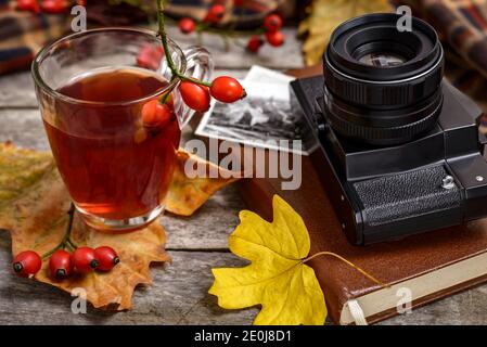 Eine Tasse Hagebuttentee mit frischen Hagebutten, Vintage-Kamera, alten Fotografien, Buch und Herbstblättern auf Holztisch. Herbstkonzept, selektiver Fokus. Stockfoto