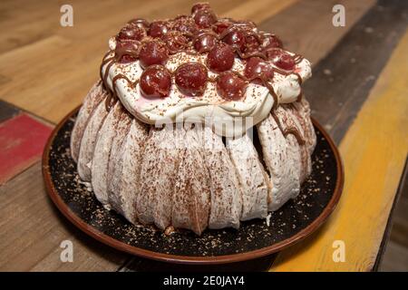 Eine köstliche pavlova-Wüste mit Sahne und Kirschen und Schokolade Sirup auf einem braunen Teller auf einer hölzernen Küchenarbeit Oben Stockfoto