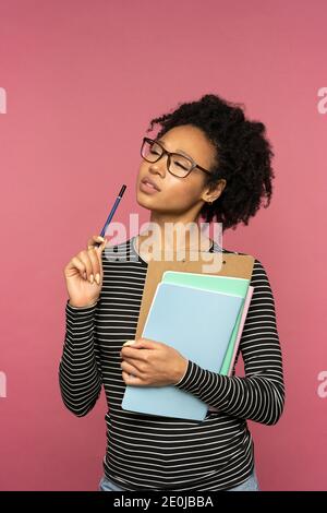 Nachdenkliche afro-amerikanische Lehrerin Frau isoliert auf rosa Studiowand. Student Mädchen tragen Brille halten Ordner, Notebooks und Stift, Denken, wegschauen. Stockfoto