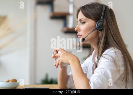 Nachdenkliche Angestellte, die auf dem Tisch sitzt, Webinar schaut, Kopfhörer trägt, mit dem Kunden per Videocomputeranruf kommuniziert, Tee trinkt Stockfoto