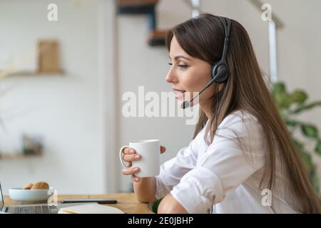 Geschäftsfrau sitzt auf dem Tisch, nimmt an Videokonferenzen, tragen Kopfhörer, Kommunikation mit dem Kunden über Video-Computer-Anruf, Tee trinken. Stockfoto