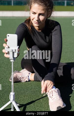 Frau in Sportswear Vlogger sitzt auf Kunstrasen im Freien, demonstriert Übungen für ihren Online-Blog, Beginn der Aufnahme auf Smartphone-Kamera o Stockfoto
