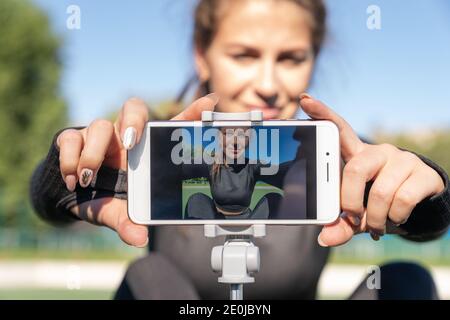 Frau in Sportswear Vlogger sitzt auf Kunstrasen im Freien, demonstriert Übungen für ihren Online-Blog, Aufzeichnungen auf Smartphone-Kamera auf Stativ. B Stockfoto