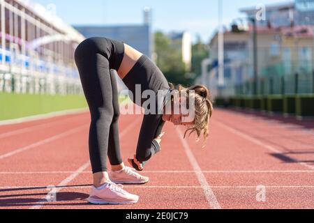 Fit junge Frau in schwarzer Sportbekleidung trainieren auf einem Laufband Gummi Stadion an sonnigen Sommertag, dabei stehen nach vorne falten Haltung, uttanasana pos Stockfoto