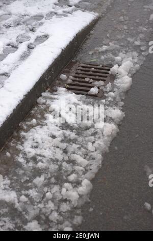 6 - Auftauen Schnee fließt in eine Metall-Bordwand Abfluss auf der Oberfläche einer asphaltierten Straße. Track sichtbar, wo ein Auto durch den Schnee gefahren hat. Stockfoto