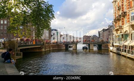 Malerische Lage an einem Kanal in Amsterdam mit einem Waterfront Cafe, Brücke mit Fahrrädern und Blumen, ein Boot und ein junges Paar genießt die Aussicht Stockfoto