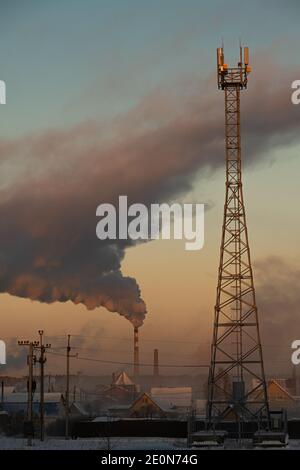 Rauch steigt aus dem Kamin des Kesselraums über der Stadt auf und der Zellenturm ist im Vordergrund. Russland, Nowosibirsk Region, Lufttemperatur -41 Stockfoto