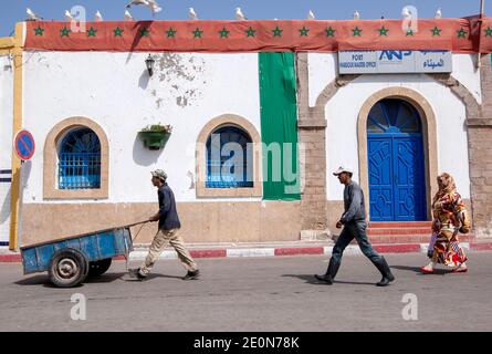 Arbeiter gehen an einer Straße am Fischereihafen von Essaouira an der Atlantikstadt in Marokko entlang, während Möwen auf dem Dach eines Gebäudes säumen. Stockfoto