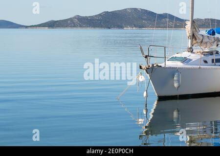 Kleines weißes Segelboot in der Adria mit der Reflexion auf der Wasseroberfläche verankert, Insel Pasman im Hintergrund, Zadar County, Kroatien Stockfoto