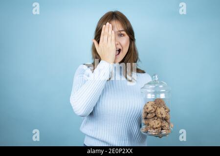 Junge schöne Frau hält Schokolade Chips Cookies Glas über isolierten blauen Hintergrund bedeckt ein Auge mit der Hand, selbstbewusstes Lächeln auf Gesicht und Surpri Stockfoto